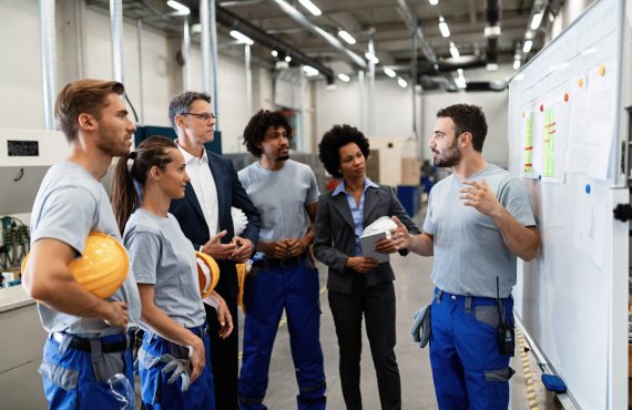 Young factory worker giving presentation of the results of the production development. Manual worker communicating with company leaders and his coworkers during business presentation in a factory.